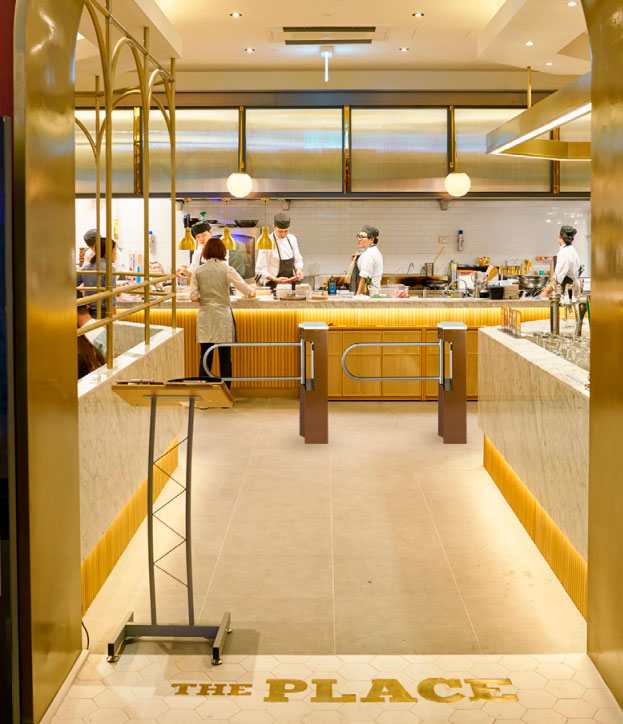 Two turnstiles distinguishing lane entrances at a food buffet, behind which a woman is standing and ordering food from several chefs behind a counter.