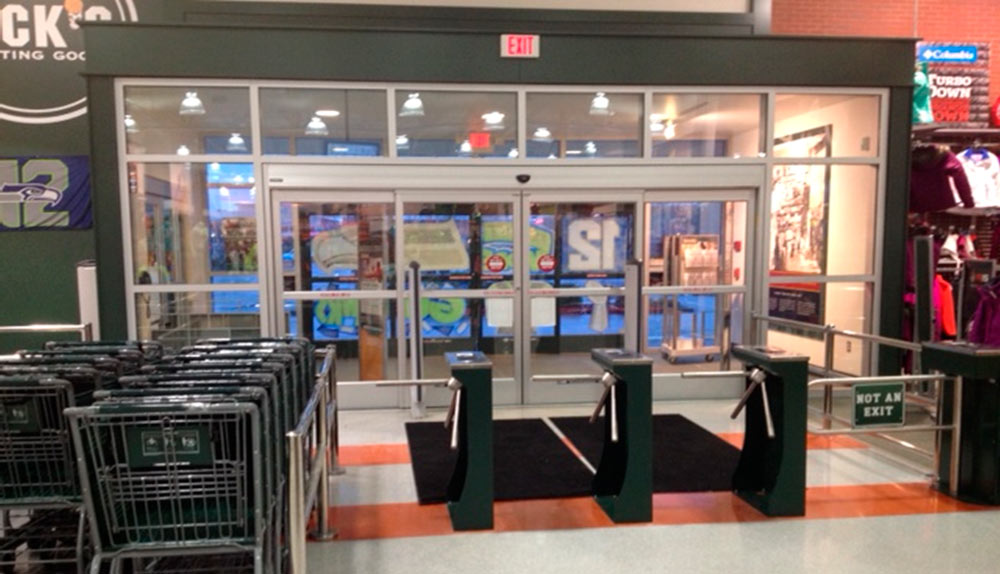 Controlled Access turnstiles in front of automatic glass entrance doors at a sporting goods store. Several shopping carts are lined up next to the three turnstiles.